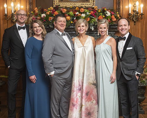 Three couples stand in front of a paneled wall and fireplace and smile at the camera